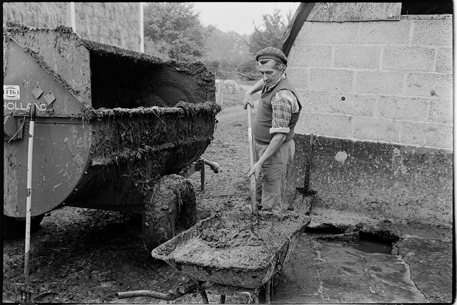 Farmer washing out milking parlour after milking and taking muck out in wheelbarrow. 
[William Medland shovelling muck from a wheelbarrow into a muck spreader in the farmyard at Hallcourt, Petrockstowe.]