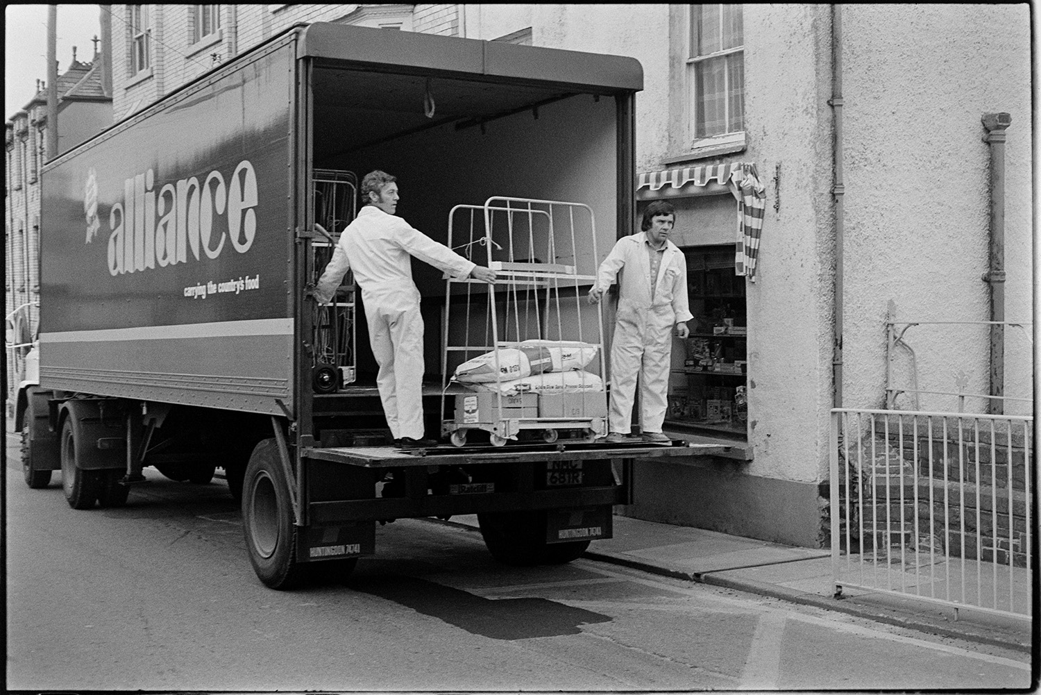 Men unloading food, groceries from lorry with tail hoist. 
[Two men unloading groceries from a lorry outside a shop in Torrington.]