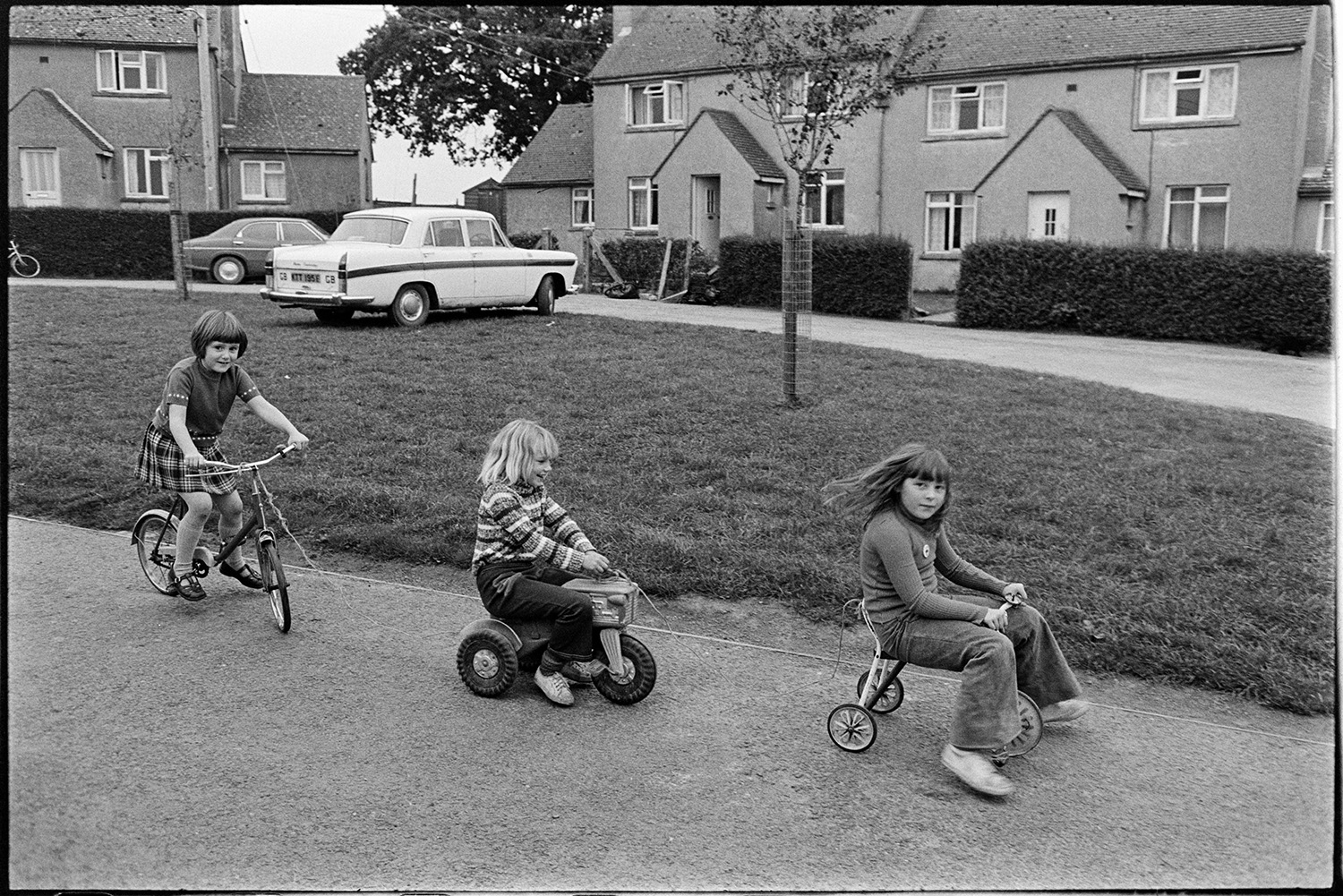 Children, girls on bicycle and tricycle on council estate. 
[Three girls riding tricycles and a bicycle on a street in front of houses in Merton.]