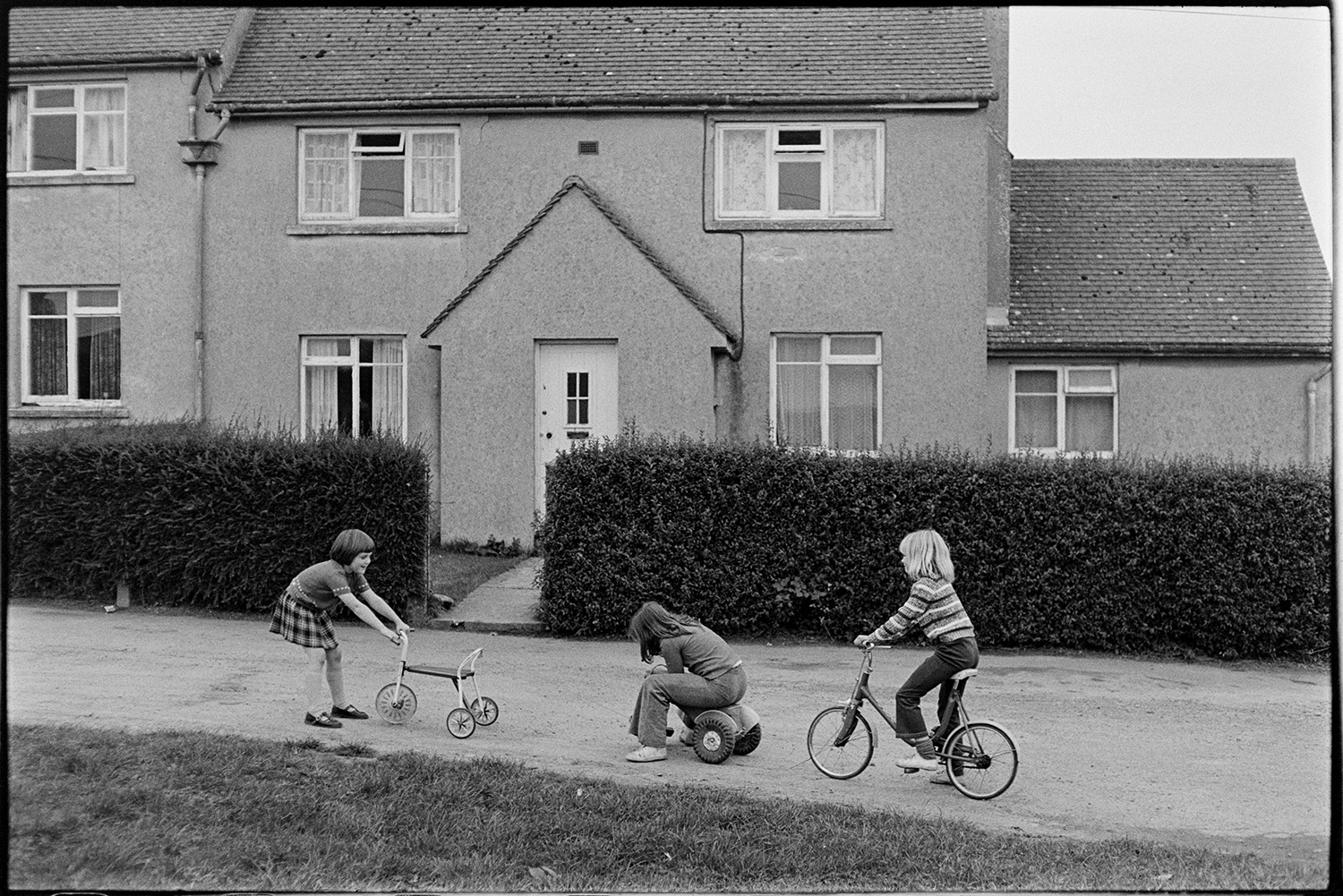 Children playing in street bicycles, tricycles. 
[Three girls playing an riding on tricycles and a bicycle on a street outside a house in Merton.]