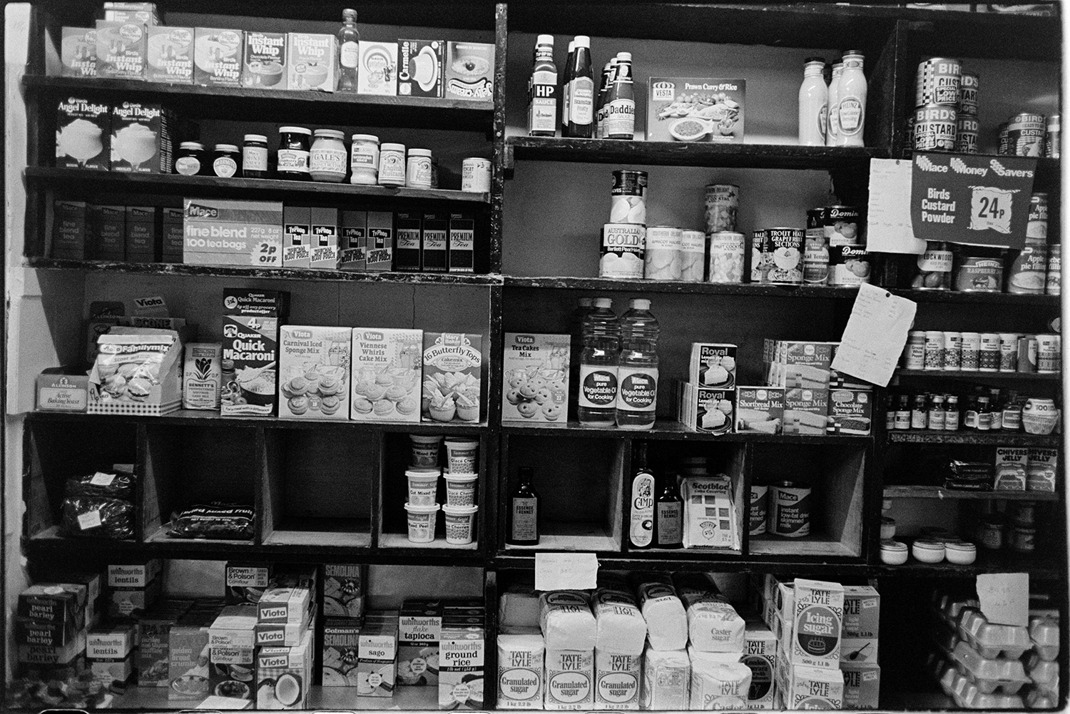 Interior of village shop with woman serving customers, goods on shelves, bacon slicer. 
[Various goods on display on the shelves in Merton Stores, including angel delight, cake mixes, HP sauce, eggs and bags of sugar.]