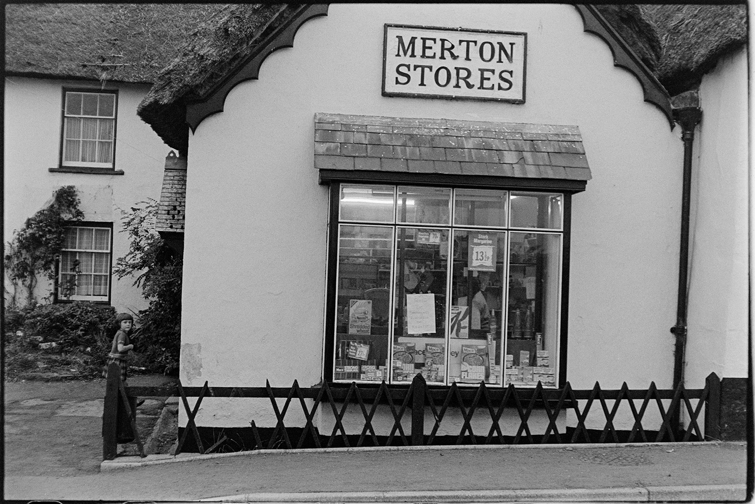 Front of village stores, shop. 
[The shop front window of Merton Stores with a wooden fence outside. A child is walking into the shop.]