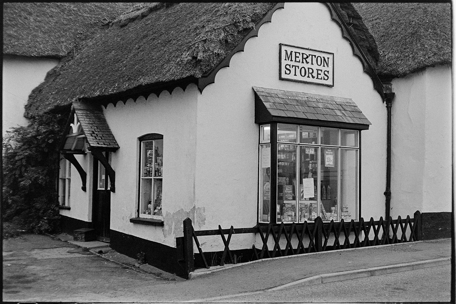 Front of village stores, shop. 
[The shop front of Merton Stores. The roof is thatched and a small porch is over the doorway. Goods are displayed in the window.]