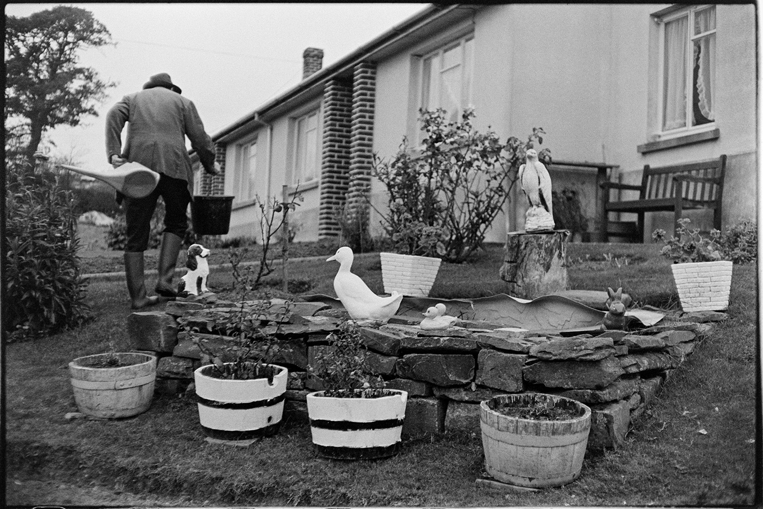 Ornamental garden with duck and dog, real dogs too! 
[Sidney Bryant holding a bucket and watering can in his garden in Merton. In the foreground is a small walled pond with garden ornaments, including a duck, squirrel and dog, and plants in pots of barrels.]