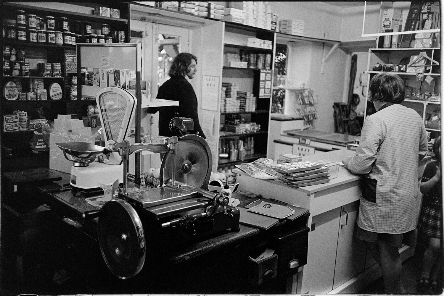 Interior of village shop with woman serving customers, goods on shelves, bacon slicer. 
[Christine Keogh, proprietor of Merton Stores, serving a customer in the shop. Jars and tins of food can be seen on the shop shelves and a bacon slicer and set of scales is on the counter.]