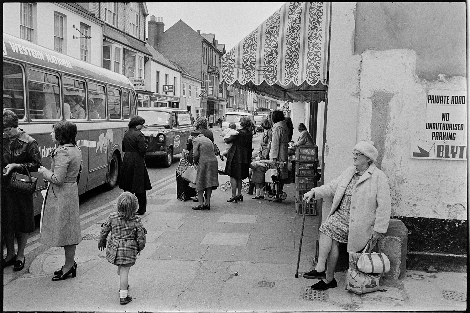 Street scene woman resting and passing bus. 
[A woman resting on a bollard by a shop front in Barnstaple High Street. Her shopping bags are by her side. Women and children are waiting in the street for a bus which has just arrived. Shop fronts are visible along the street, including one in the foreground with an awning and crates outside.]