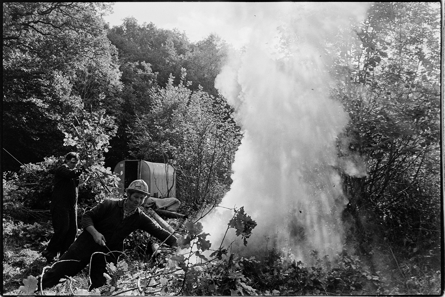 Men burning hedge cuttings during bridge renovation. Bonfire. 
[Two men burning hedge trimmings on a bonfire at Addisford, Dolton. Trees and a corrugated iron shed can be seen in the background.]