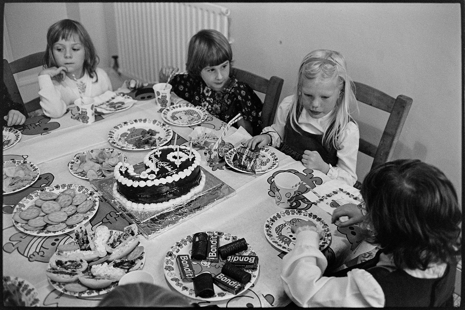 Birthday tea party, wonderful spread with cakes and children stuffing themselves, eating. 
[Girls sat round a table eating party food at the 6th birthday of Hilary and Roger Hill's child, at Marwood House. The table is laid with chocolates, cakes, biscuits and crisps, with a birthday cake in the shape of the number 6 in the middle.]