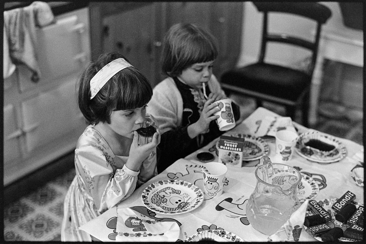 Birthday tea party, wonderful spread with cakes and children stuffing themselves, eating. 
[Two children sat at a table eating and drinking at the 6th birthday of Hilary and Roger Hill's child, at Marwood House.  The table is laid with Mr Men themed plates and table cloth. A jug of juice and chocolates can also be seen on the table.]