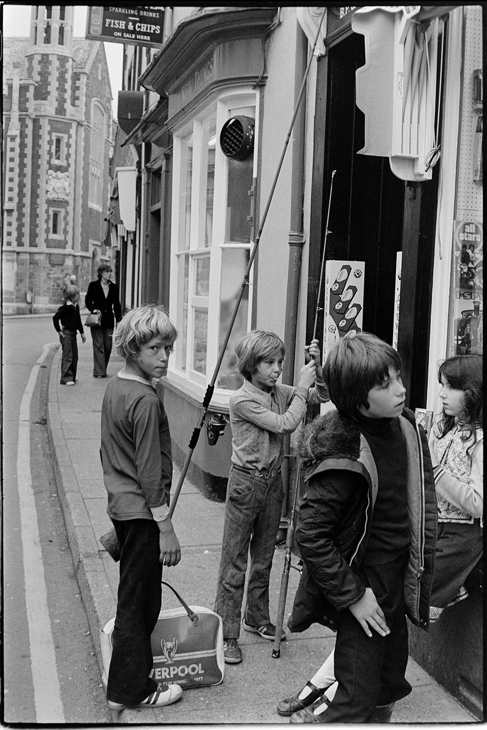 Shops and stalls at Pannier Market, WI Stall. 
[Children outside a shop front on a street in Bideford. Two of the children are holding fishing rods. A fish and chip shop sign can be seen in the background.]