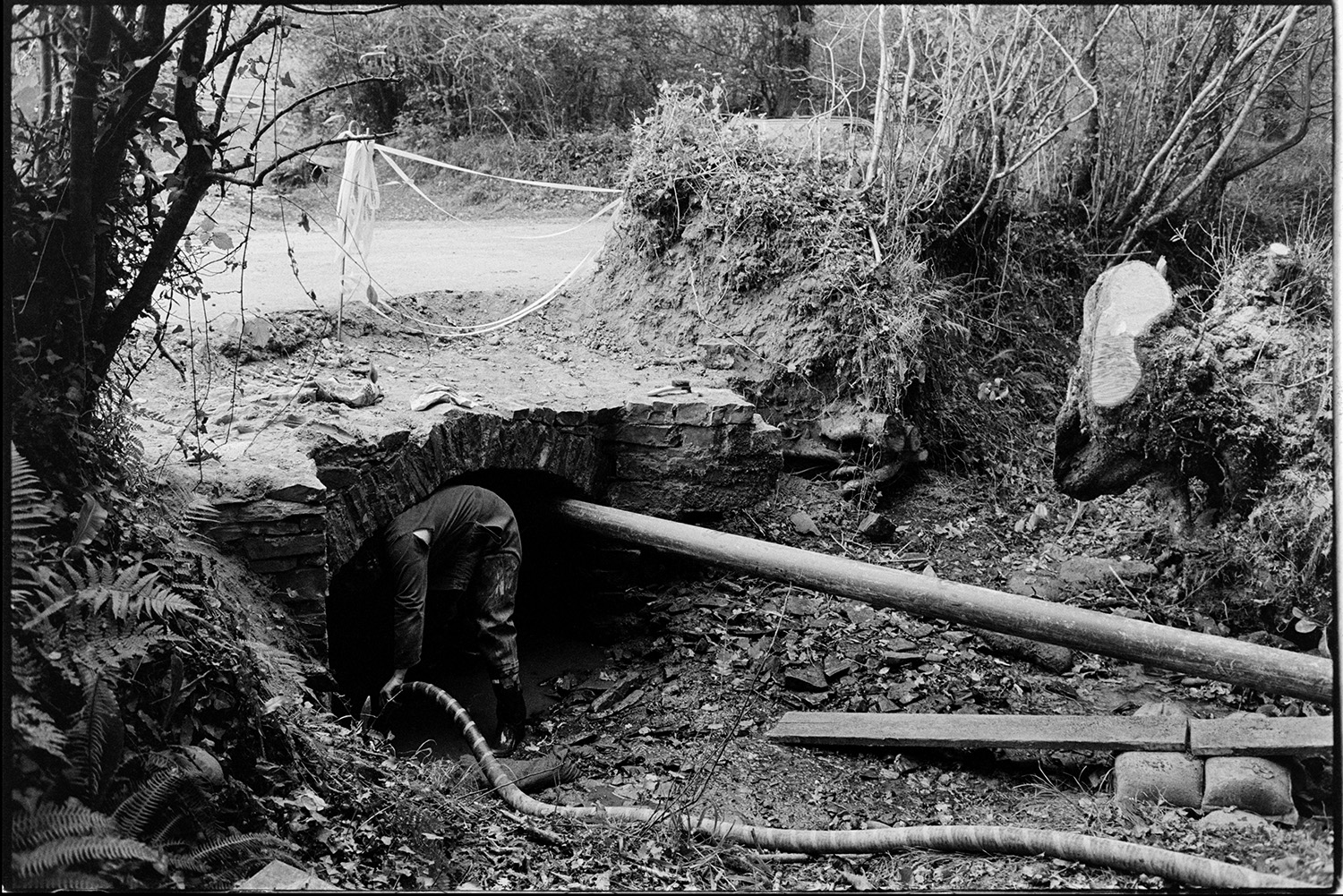 Men repairing bridge, crawling under road. 
[Man repairing a bridge at Addisford, Dolton. He is crawling under the arch of the bridge.]