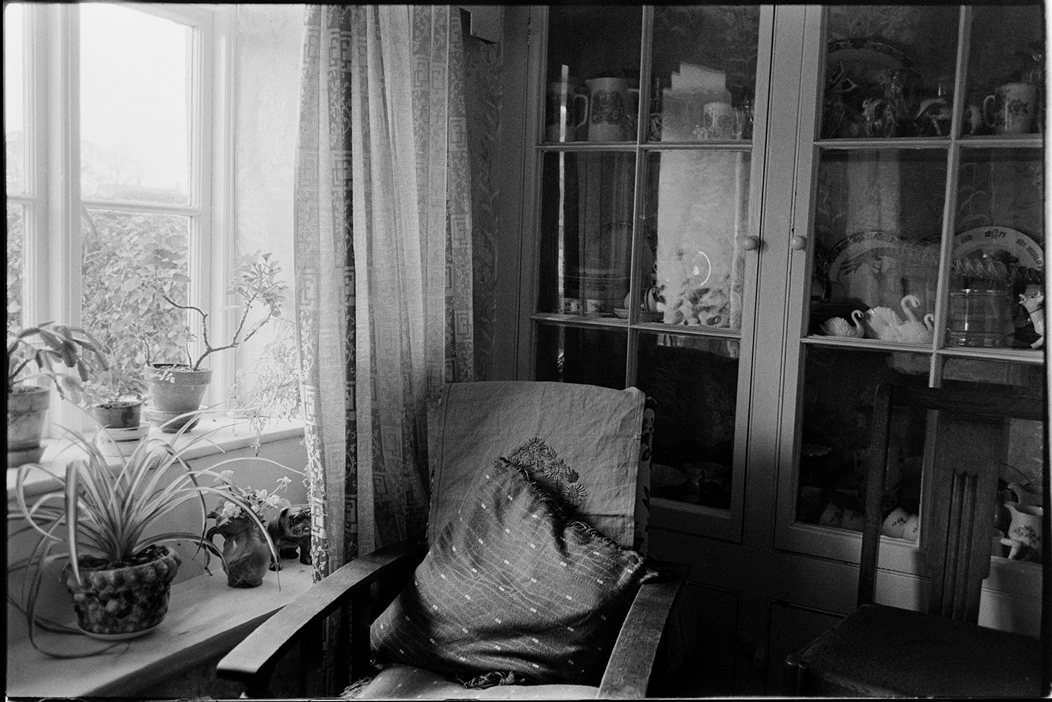 Interior of room with dresser and ornaments, corn dolly. 
[A dresser behind a chair in a room. The dresser is filled with china and ornaments. Pot plants are visible in the window sill.]