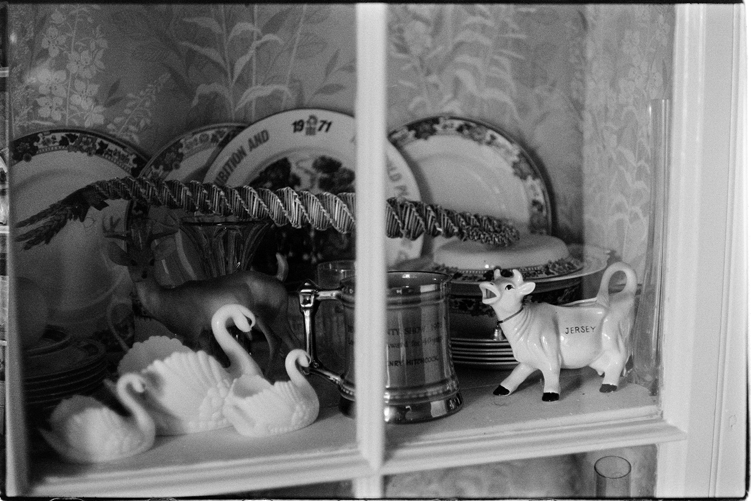 Interior of room with dresser and ornaments, corn dolly. 
[A close up view of china plates, china ornaments, including a cow and swans, and a silver tankard in a dresser. A corn dolly is resting on top of the ornaments.]
