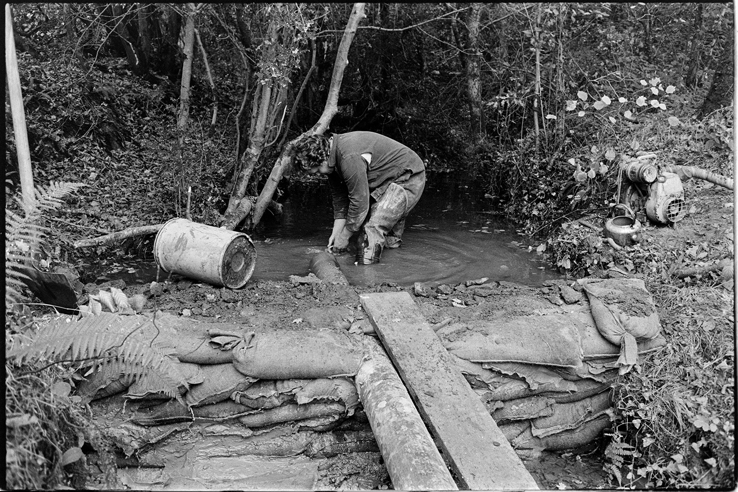 Men repairing bridge, crawling under road. 
[A man repairing a bridge at Addisford, Dolton. He is checking the pipe which is pumping water from the stream away from the bridge, behind a wall of sand bags.]