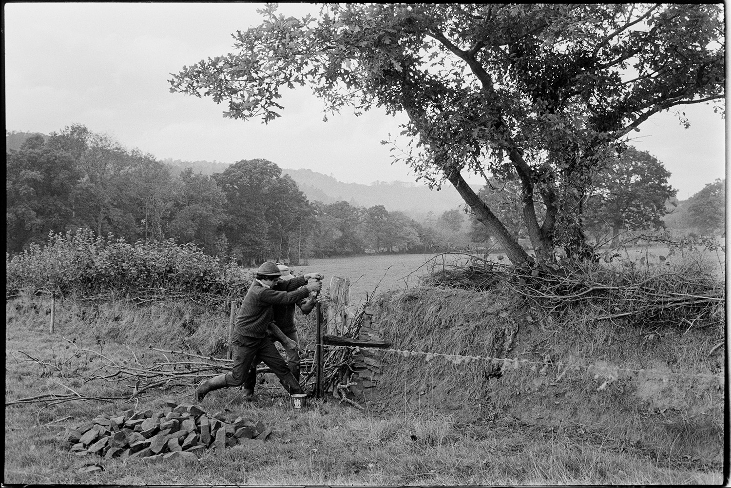Farmers mending fencing. 
[Alf Pugsley and Stephen Squire mending a barb wire fence by a hedge in a field at Lower Langham, Dolton. A pile of stones and branches are nearby in the field.]