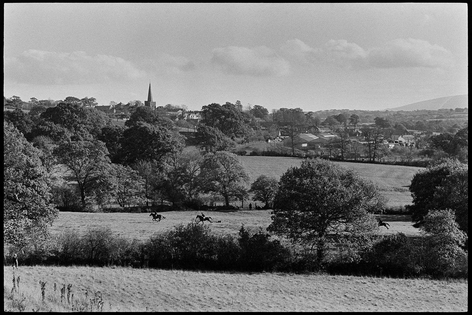 Hunt followers. 
[Huntsmen on horseback riding through a field in Hatherleigh. The village and church spire can be seen behind trees in the background.]