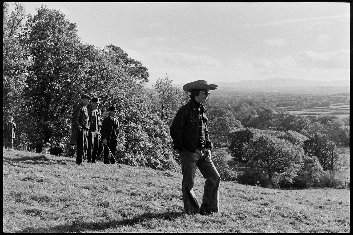 Hunt followers. 
[Men in a field watching a hunt in Hatherleigh. A man in the foreground is wearing sunglasses and a cowboy hat.]