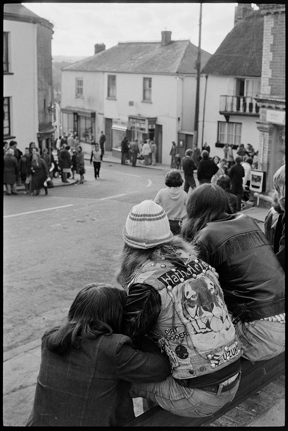 People waiting for Carnival to start, drinking and waiting in street. 
[People waiting in Market Street for Hatherleigh Carnival to start. In the foreground teenagers wearing leather and denim jackets are sat on a wall. One of the jackets has a picture of a tiger on it.]
