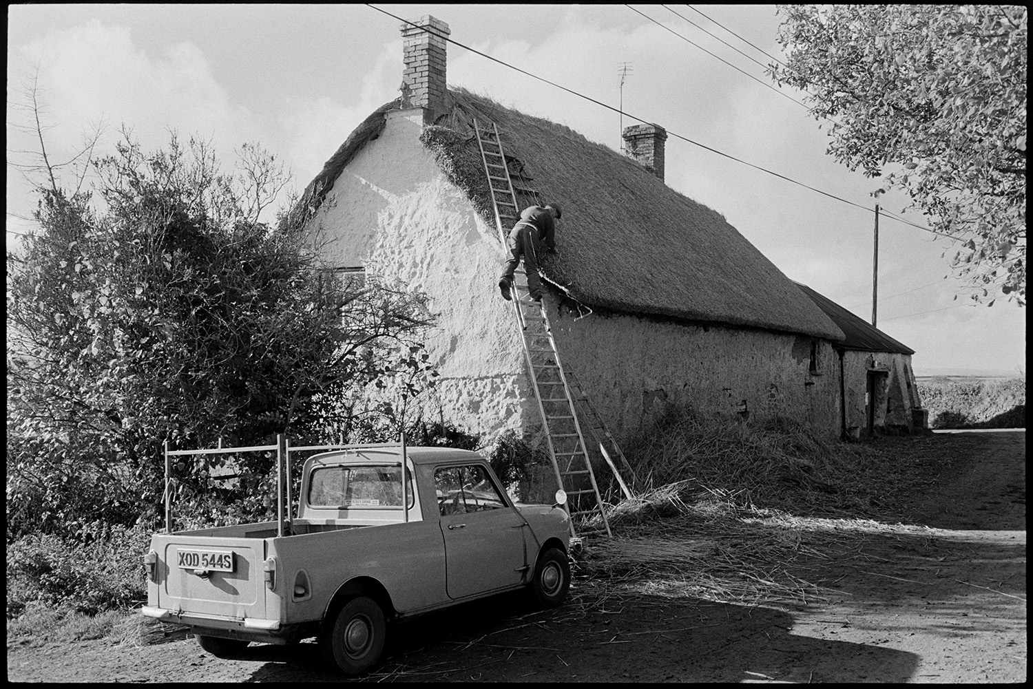 Thatcher working in village and talking to farmer. 
[Stanley Cole up a ladder thatching a cottage roof in Upcott, Dolton. A pile of reed is by the house wall and a truck is parked on the road.]