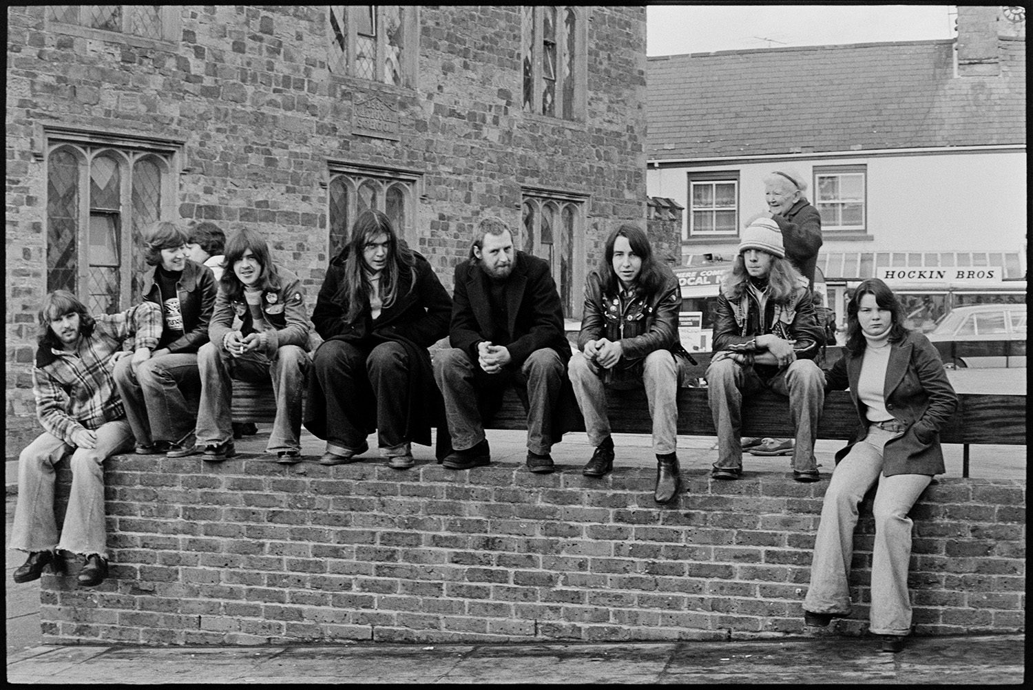 Woman and young men waiting for carnival. 
[A young woman and young men sat on a wall waiting for Hatherleigh Carnival. Hockin Brothers shop front can be seen in the background.]