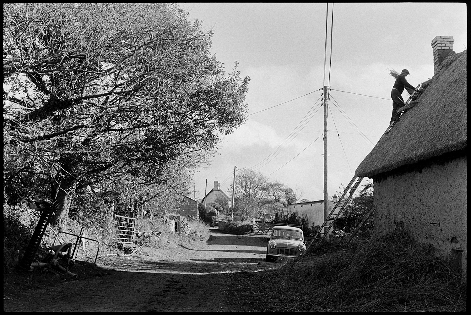 Thatcher working in village and talking to farmer. 
[Stanley Cole up a ladder thatching a cottage roof in Upcott, Dolton. A pile of reed is by the house wall. A truck is parked in the road by the house. A field gate and various items of farm machinery can be seen on the opposite side of the road.]