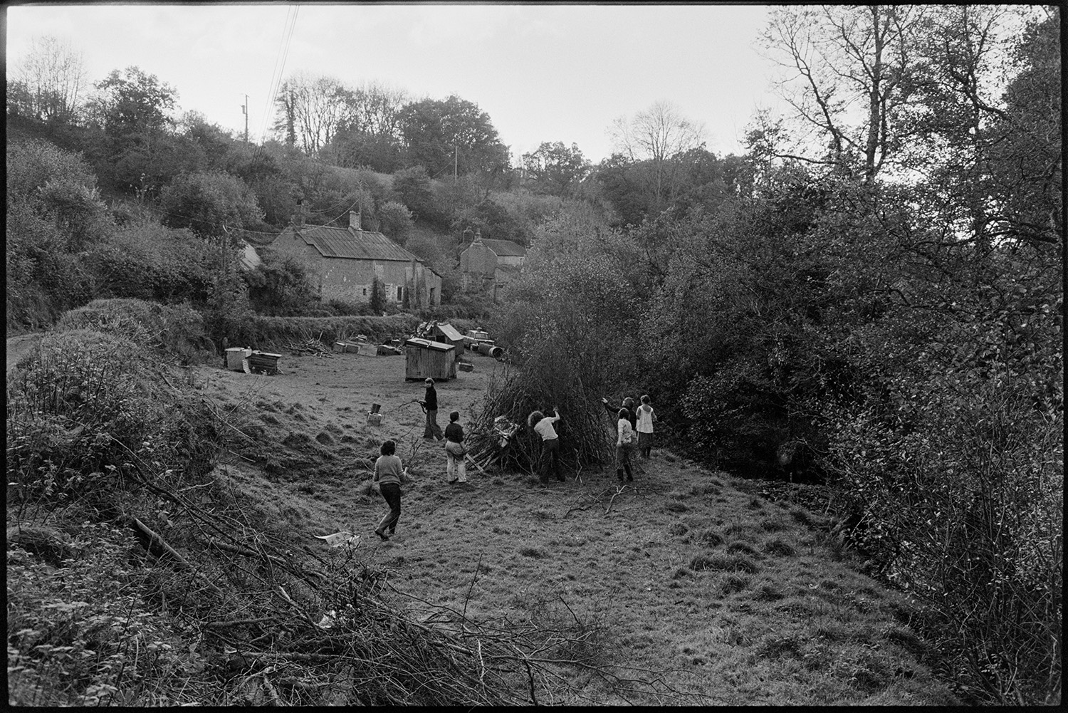 People making bonfire for Guy Fawkes night. 
[Men and women gathering branches and making a bonfire in a field at Millhams, Dolton. Sheds and cottages can be seen in the background.]