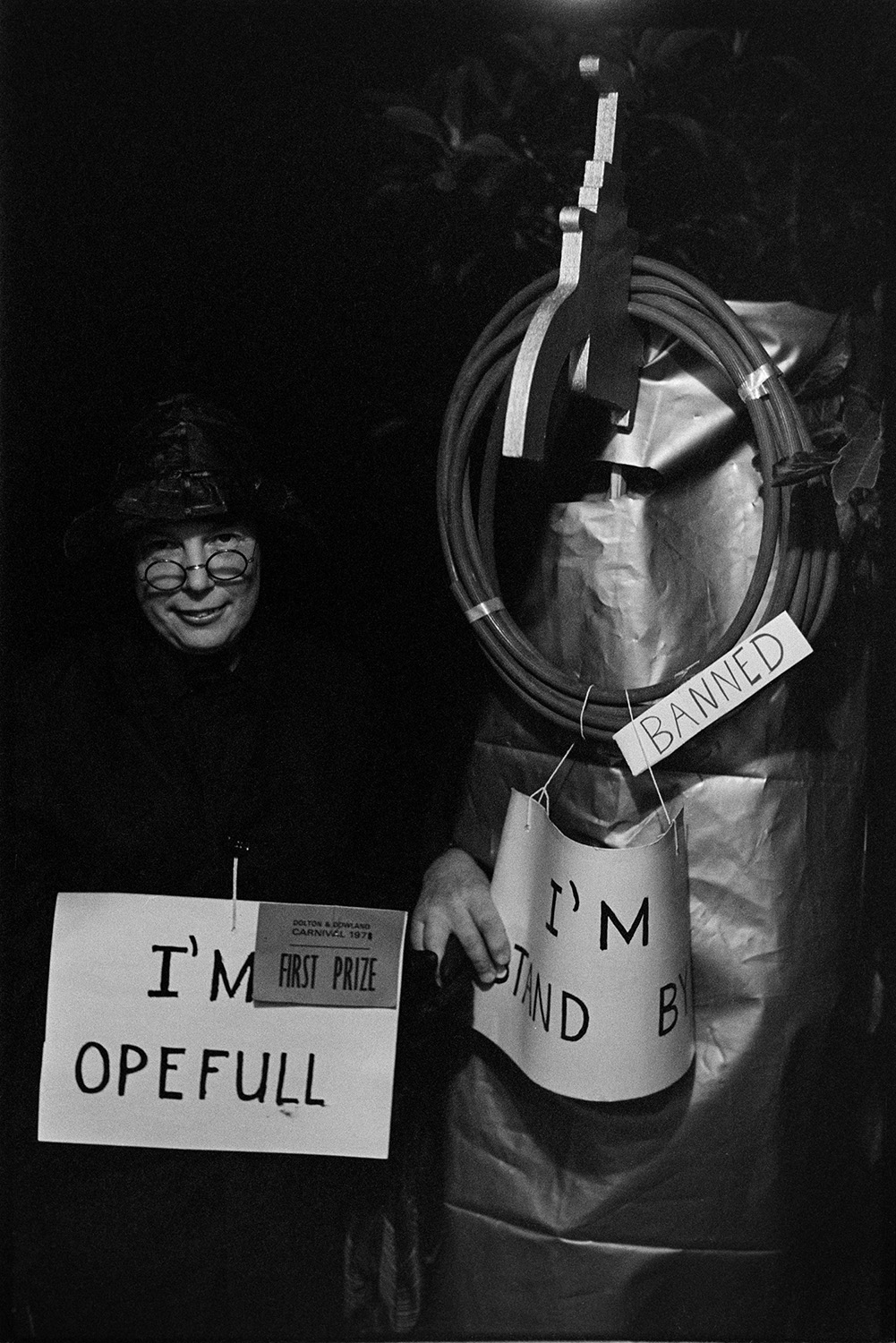 Stalls and floats at carnival at night, Carnival Queen. 
[Two people in costumes at Dolton Carnival who won first prize. One person is carrying an 'I'm Opefull' sign and the other is dressed as a tap with a pipe and is carrying a sign reading 'I'm Stand By'.]