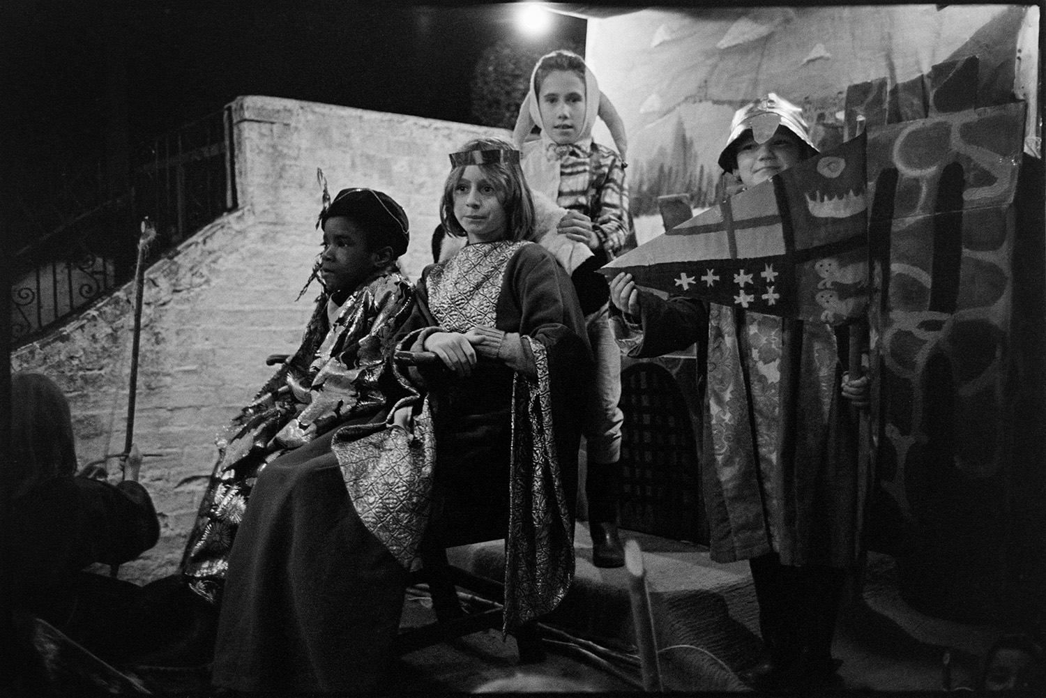 Carnival floats at night with flares being lit. 
[Children dressed as a King, Queen, Courtier and Jester on a carnival float at Hatherleigh Carnival, at night.]