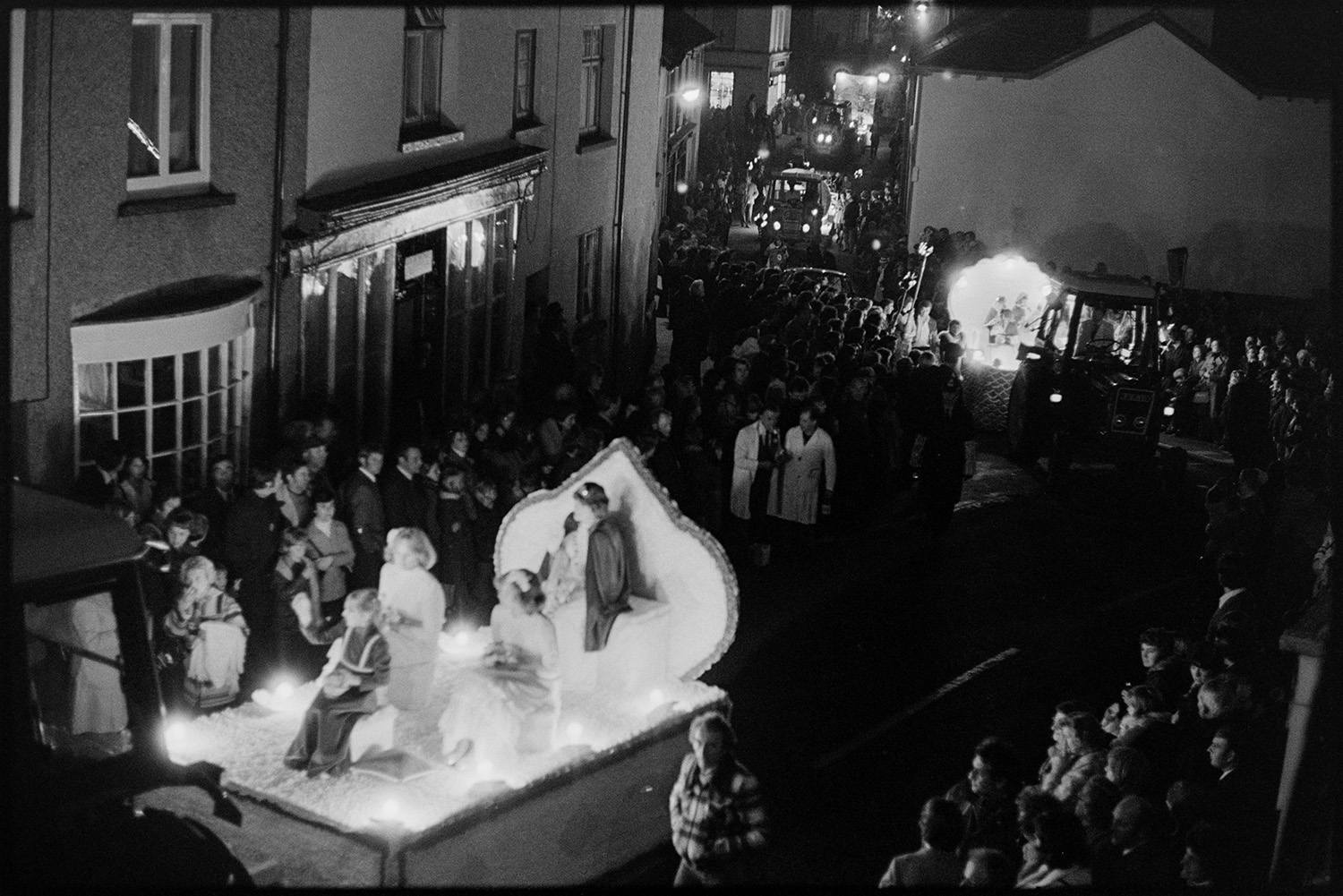 Carnival floats at night with torches. 
[Spectators watching carnival floats parade through the town for Hatherleigh Carnival, at night. A carnival float can be seen in the foreground, possibly with the Carnival Queen and her attendants.]
