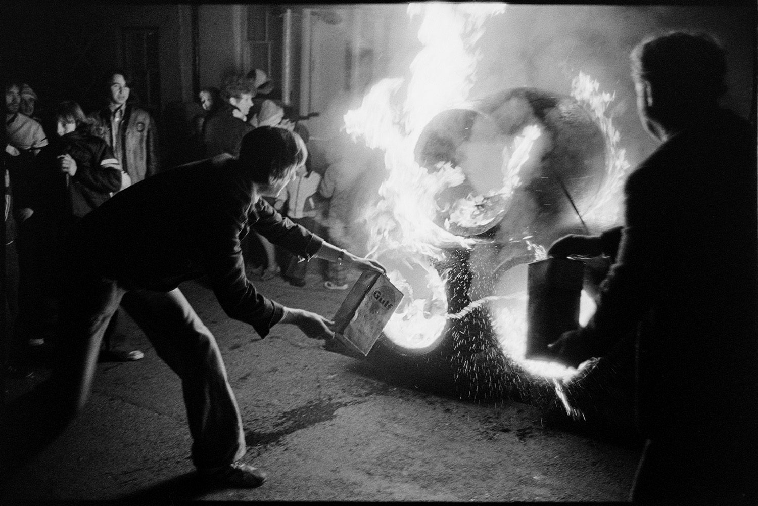 Carnival, blazing tar barrels being lit, before run round town. 
[Men throwing fuel onto alight tar barrels before they are paraded around Hatherleigh for Hatherleigh Carnival.]