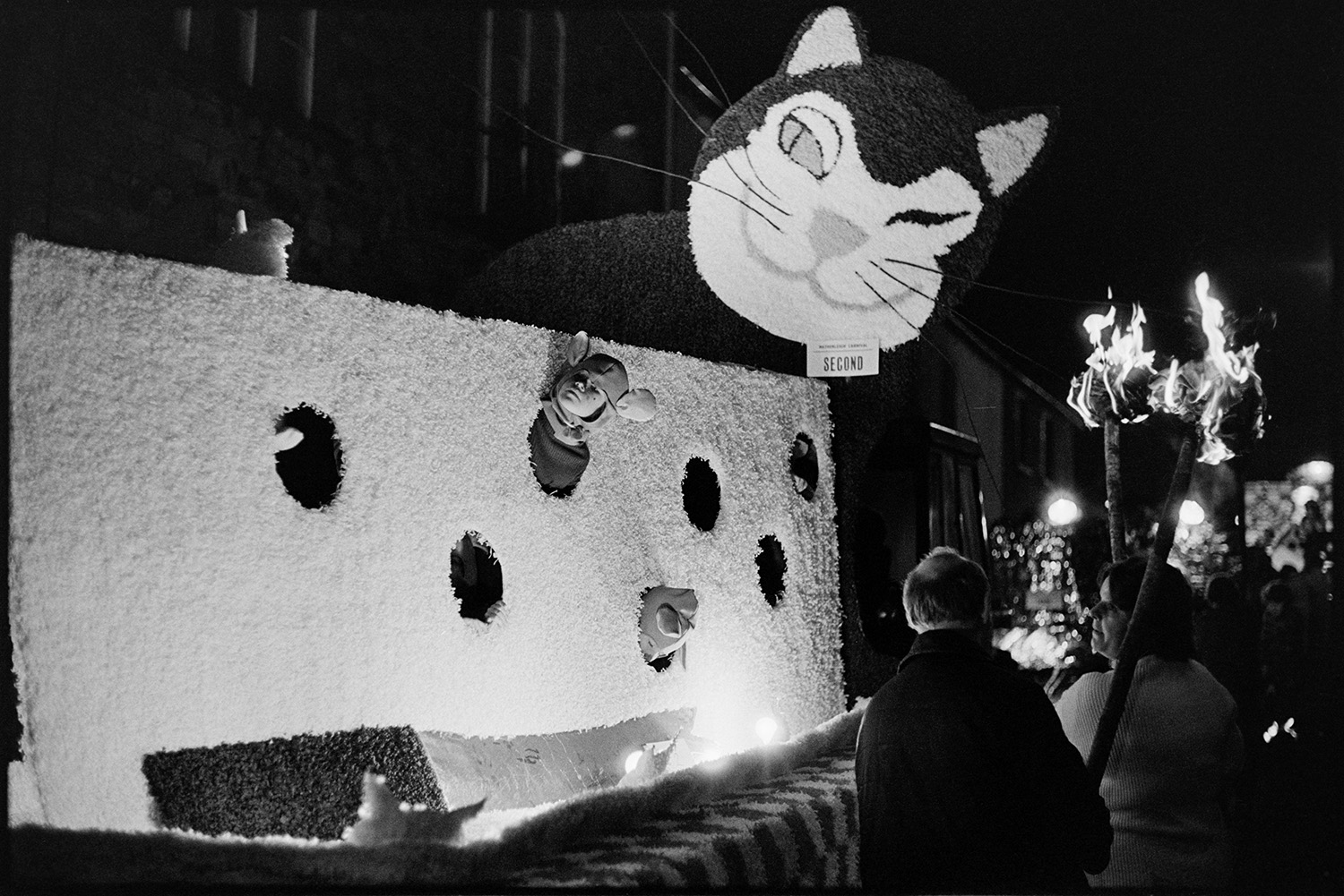 Carnival floats at night with torches. 
[A carnival float with a cat looking for mice in a piece of cheese parading through a street at Hatherleigh carnival, at night. A man and woman are walking past the float holding flaming torches. The float won second prize.]