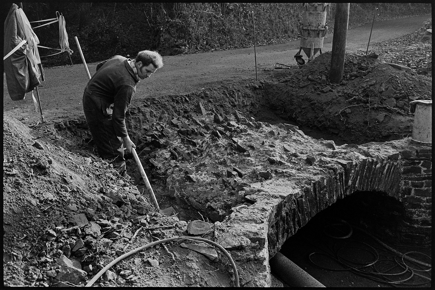 Men repairing bridge, details of construction. 
[A man repairing a bridge at Addisford, Dolton. He is digging out earth using a shovel. A road going over the top of the bridge can be seen behind him.]