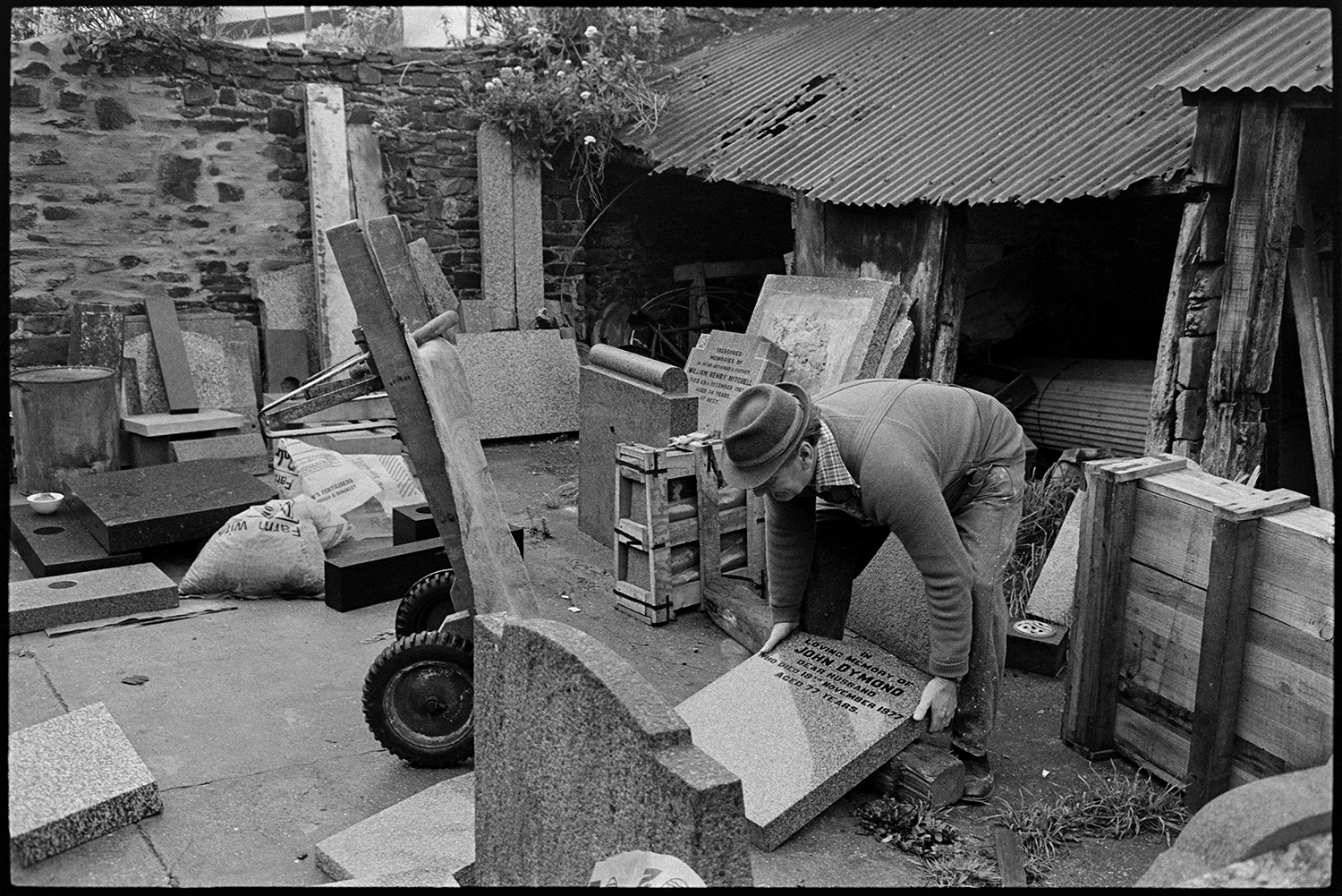 Monumental mason at work on headstone in workshop and loading stone into car. 
[Bruce Edyvean, a monumental mason, lifting a headstone for a grave onto a sack truck in the yard outside his workshop at New Street, Torrington. Other gravestones are visible in the yard.]