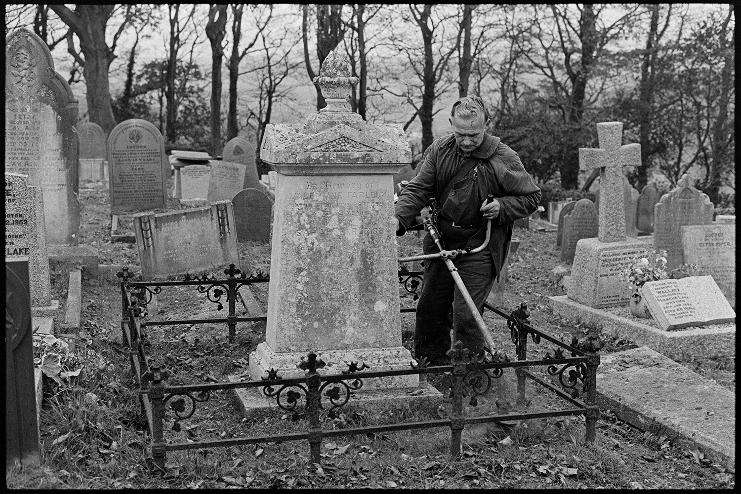 Man cutting grass round gravestones in cemetery with strimmer. 
[A man strimming grass around a gravestone with railings in Torrington Cemetery.]