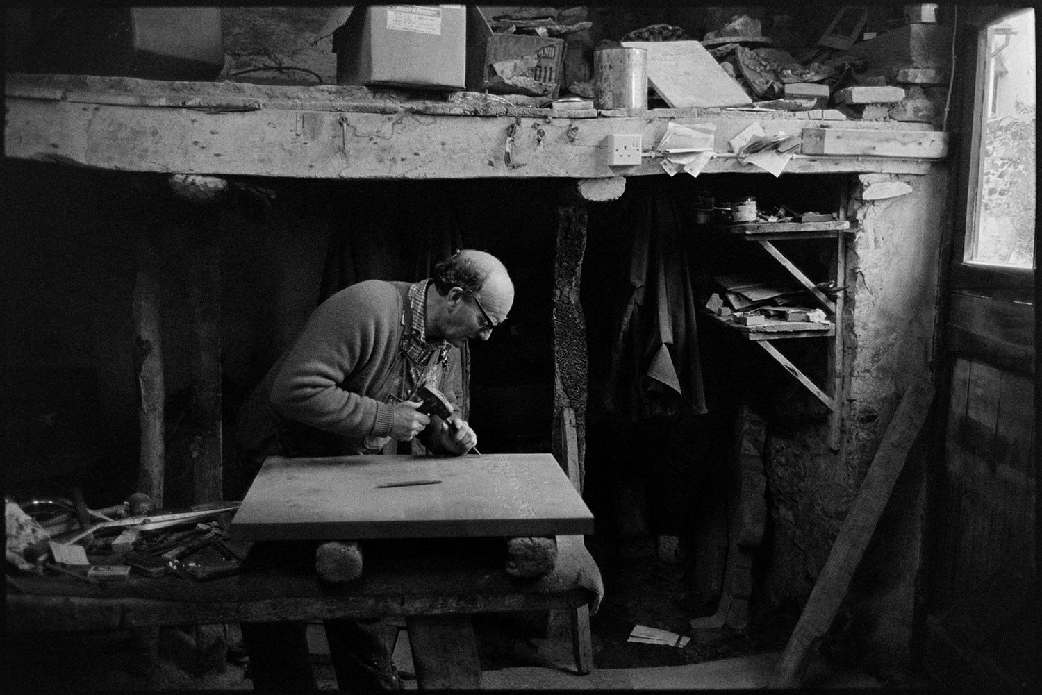 Monumental mason at work on headstone in workshop and loading stone into car. 
[Bruce Edyvean, a monumental mason, working on a headstone for a grave, in his workshop in New Street, Torrington. The gravestone is resting on a workbench and he is using a hammer and chisel to engrave the wording on the stone.]