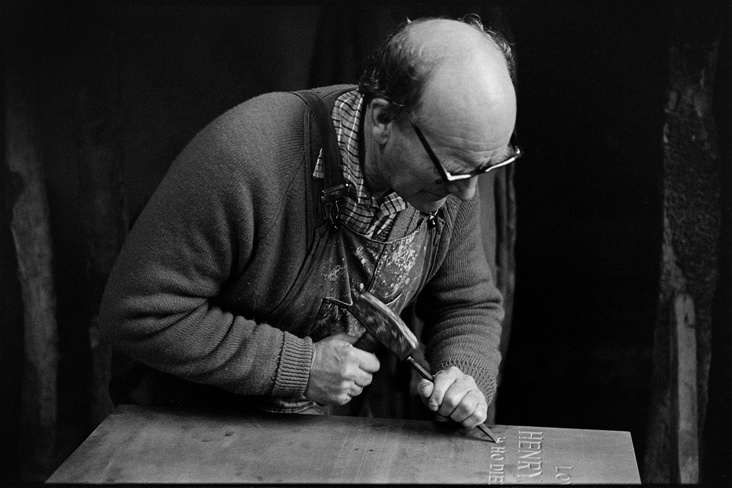 Monumental mason at work on headstone in workshop and loading stone into car. 
[Bruce Edyvean, a monumental mason, engraving a headstone for a grave in his workshop in New Street, Torrington. He is using a hammer and chisel to engrave the stone.]