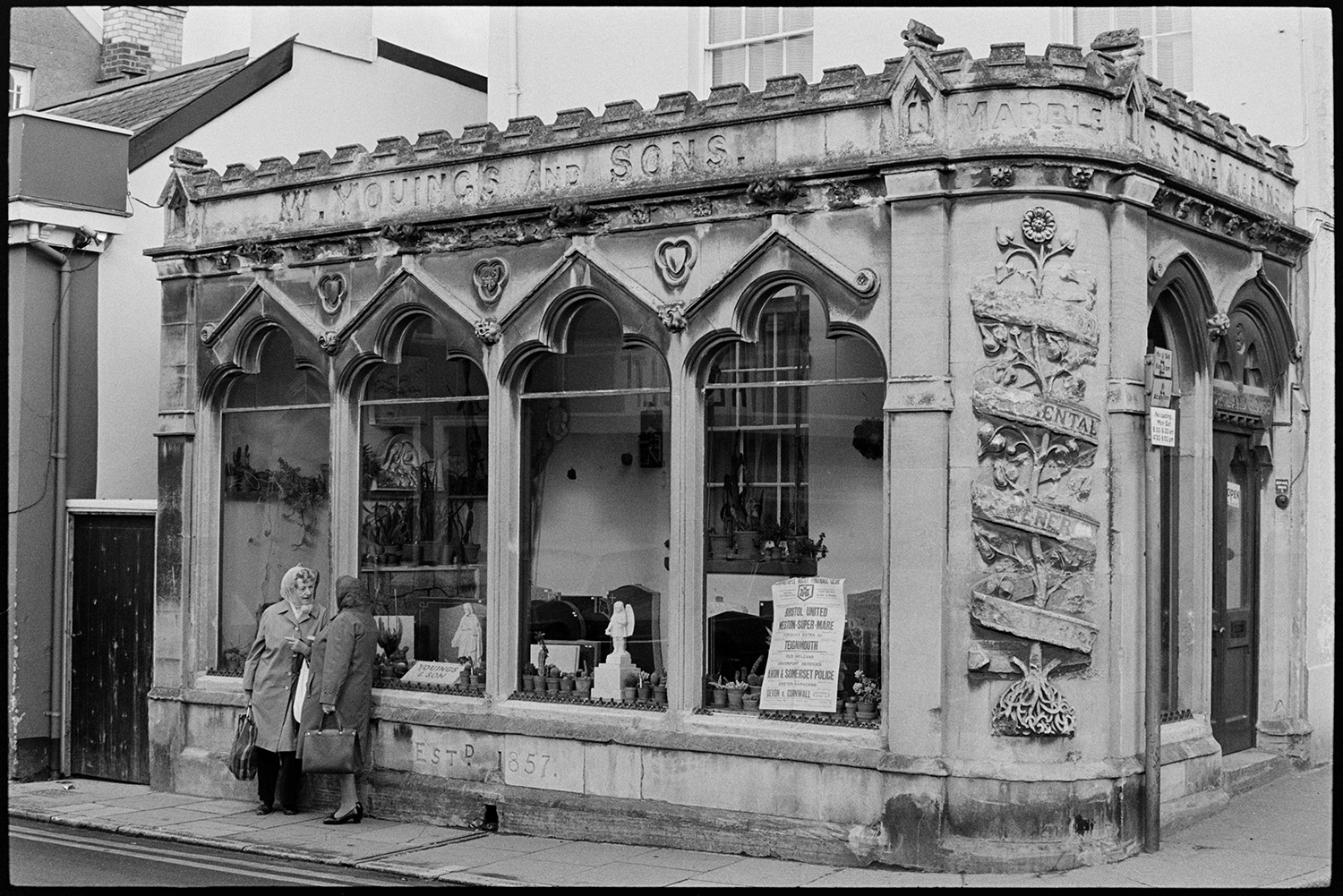 Ornately carved monumental masons premises (Victorian). 
[Two women talking outside the shop front of Youings & Son, Monumental Masons in Bear Street, Barnstaple. The shop front is ornately carved. Gravestones, statues and pot plants are visible inside the premises.]