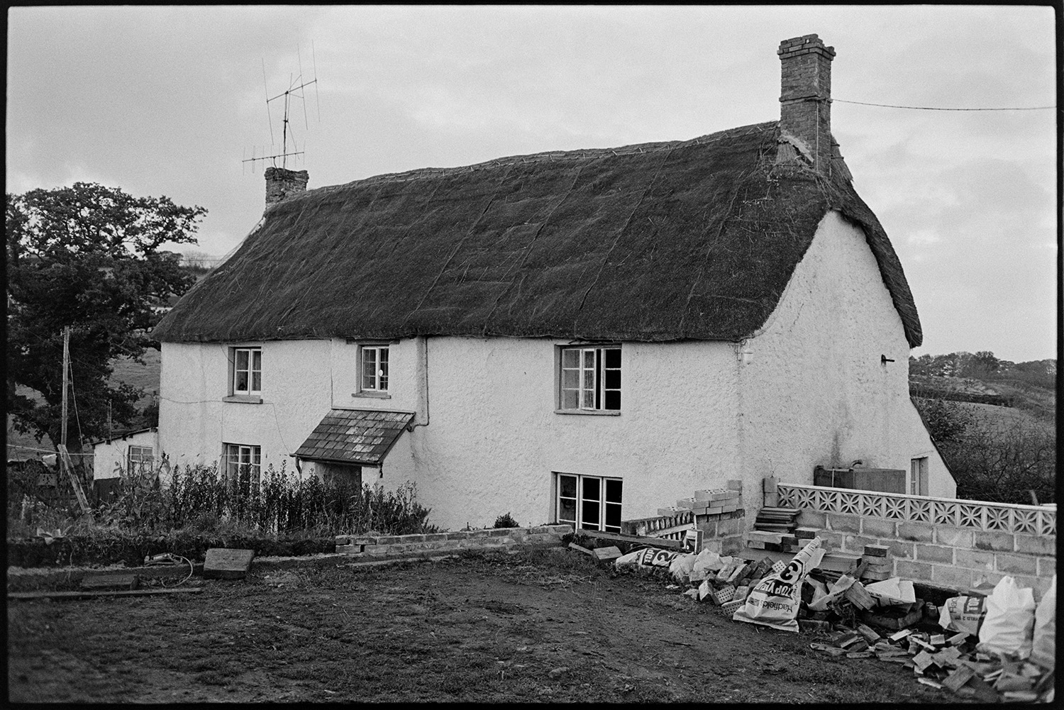 Cob and thatch farmhouse. 
[The cob and thatch farmhouse at Hallcourt, Petrockstowe. Various construction materials and a newly built wall can be seen by the side of the house.]