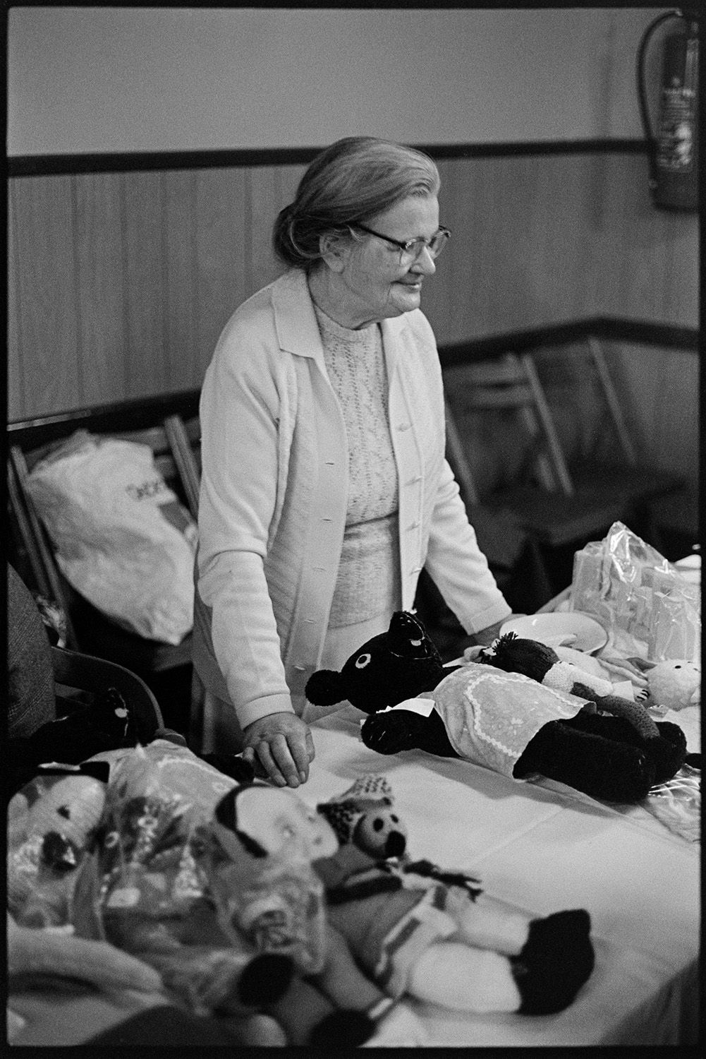 Jumble sale in village hall. Women looking through second hand clothes. 
[A woman running a stall with knitted toys and dolls at a jumble sale in Dolton Village Hall.]