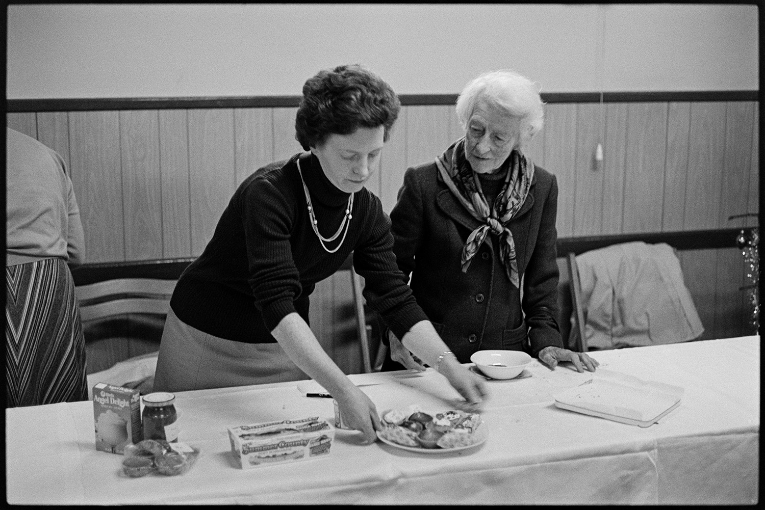 Two women at jumble sale with remains of produce. 
[Mrs Heal, on the left, and Eve Lynch-Blosse with the left overs from a cake stall at a jumble sale in Dolton Village Hall.]