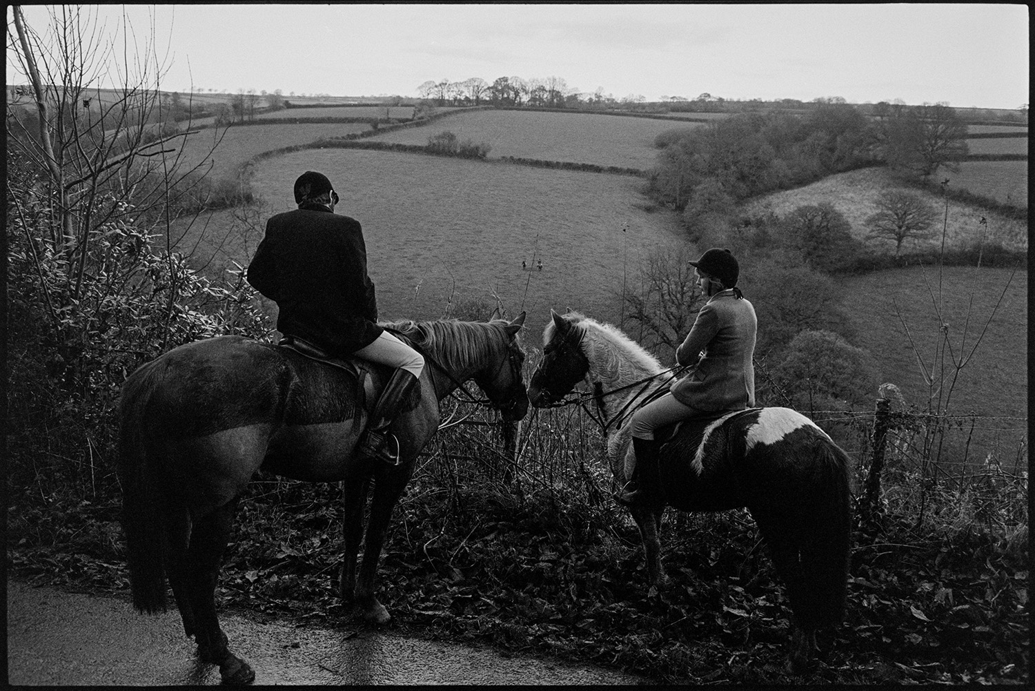 Hunters, horses and followers waiting for a scent, listening for hounds. 
[Two riders, a man and a woman, watching huntsmen in a field below, from the side of a road at Halsdon, Dolton.]