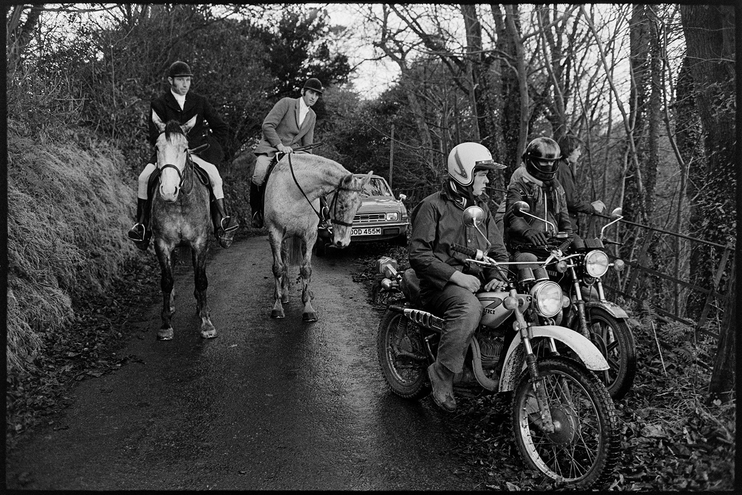 Hunters, horses and followers waiting for a scent, listening for hounds. 
[Two huntsmen in a lane at Halsdon, Dolton. They are passing hunt followers on motorbikes.]