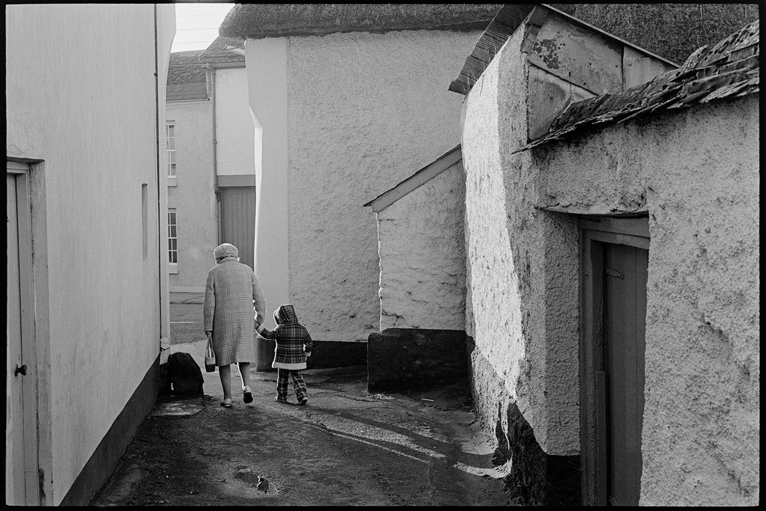 Back lane with cob walls and passers by. 
[A woman and child walking along Buddle Lane, Hatherleigh past the walls of a cob cottage.]