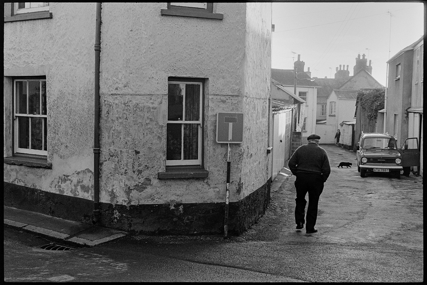 Back lane with cob walls and passers by. 
[A man walking down Buddle Lane, Hatherleigh. A parked car and a cat can be seen further down the street.]