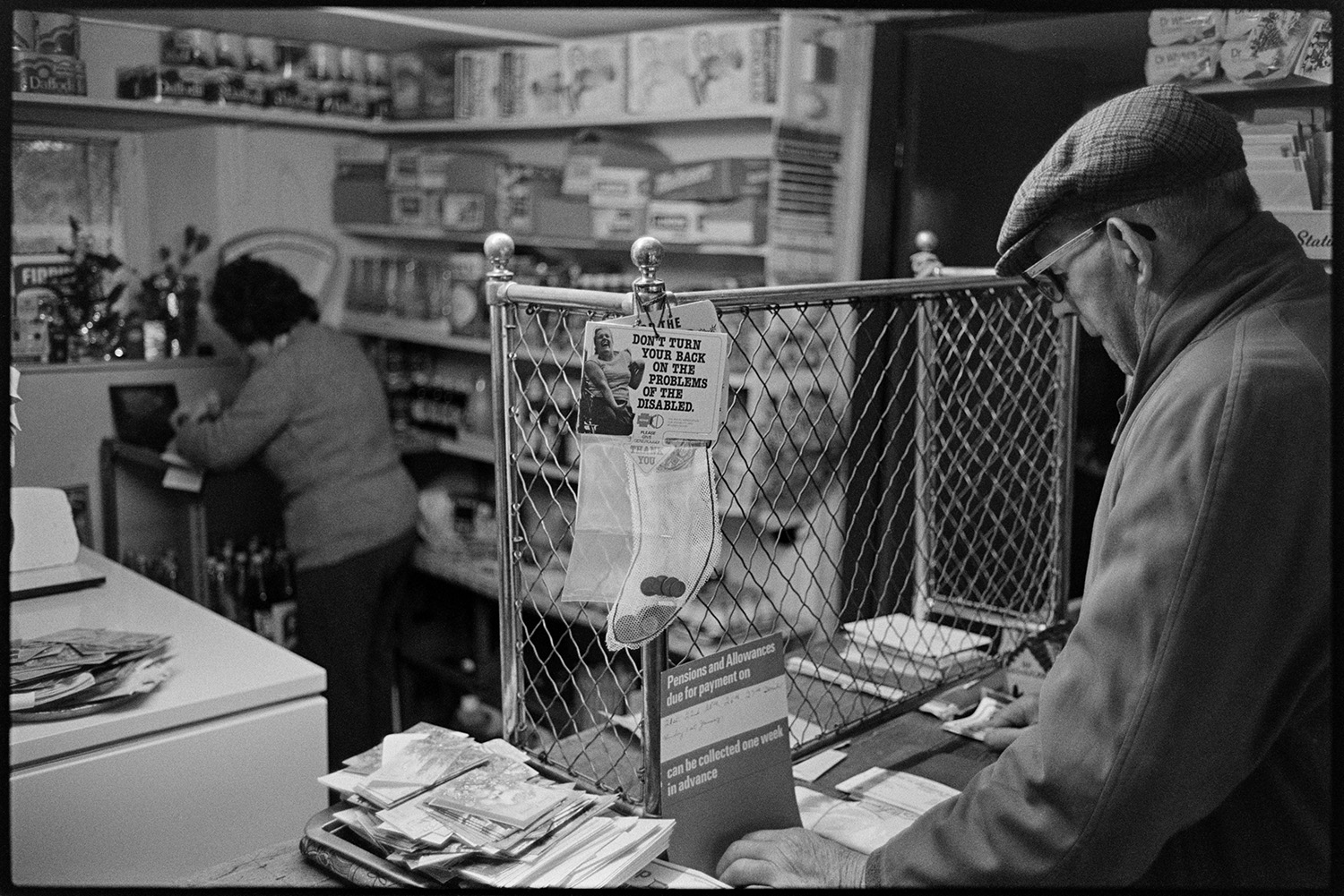 Interior, counter, Post Office Stores with woman serving customer behind old grill. 
[Enid Stentiford serving a man in Monkokehampton Post Office. She is stood by a set of scales. A mesh or grill screen is on the counter. Various cards and a poster about pensions is also on the counter.