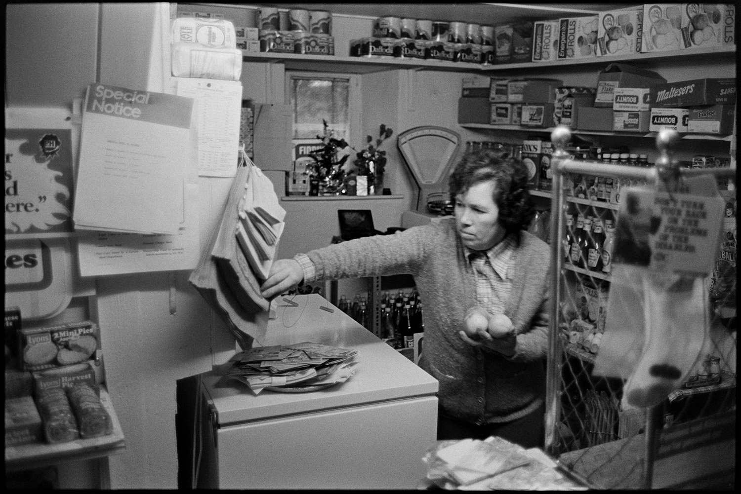 Interior, counter, Post Office Stores with woman serving customer behind old grill. 
[Enid Stentiford behind the counter of Monkokehampton Post Office. She is holding some apples which she is about to place in a paper bag. Various goods are on display on the shelves including, pies, tomato sauce and boxes of chocolate bars.]