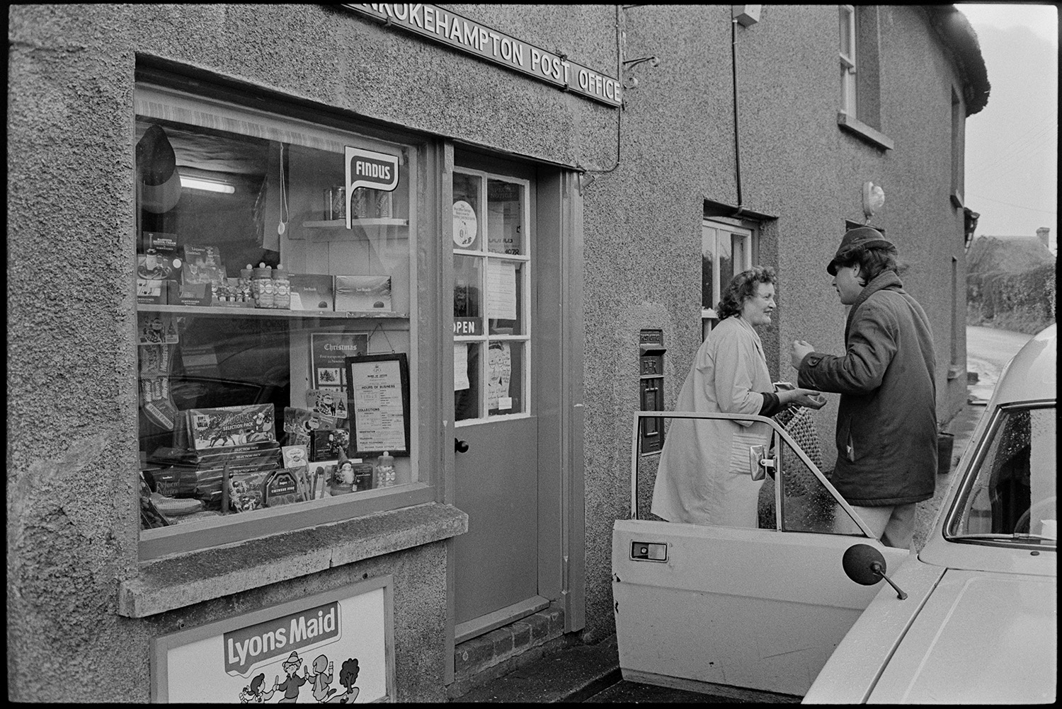 Baker delivering bread round village and outside Post Office from van. 
[A woman buying something from an A Guy & Sons Ltd of Exbourne baker's van outside Monkokehampton Post Office. Jars of sweets and boxes of chocolate can be seen in the Post Office window.]