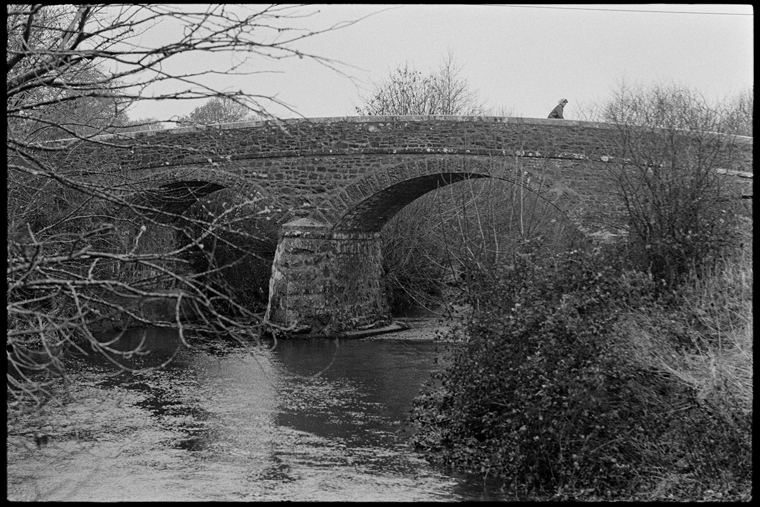 Ancient stone bridge. 
[A person walking or cycling over a stone bridge with arches at New Bridge, Monkokehampton.]