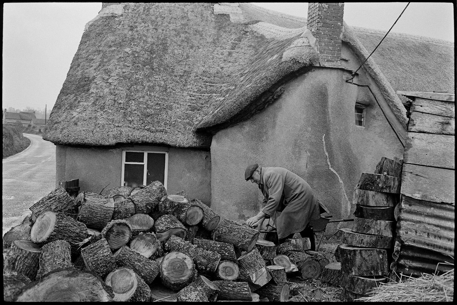 Man taking logs from woodpile in front of thatched cottage. 
[Mr Crocker gathering logs from a woodpile outside a thatched cottage in Monkokehampton.]