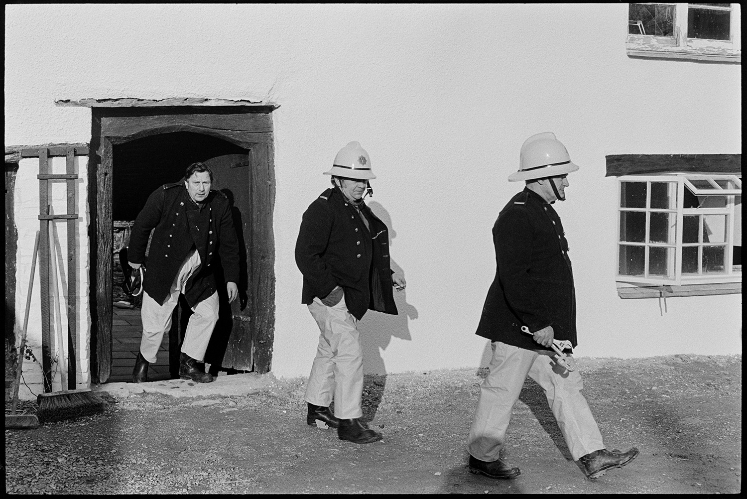 Firemen and fire engine at scene of small fire. 
[Three firemen coming out of a the entrance to a cottage after attending a small fire, at Hackwells, Dolton. Brushes are lent against the cottage wall.]